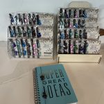 Retail countertop arrangement showcasing packaged Inspirational Book Bracelets in a tier stand and wicker basket alongside a light blue journal reading "SUPER GREAT IDEAS".