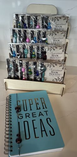 Retail countertop arrangement showcasing packaged Inspirational Book Bracelets in a tier stand and wicker basket alongside a light blue journal reading "SUPER GREAT IDEAS".